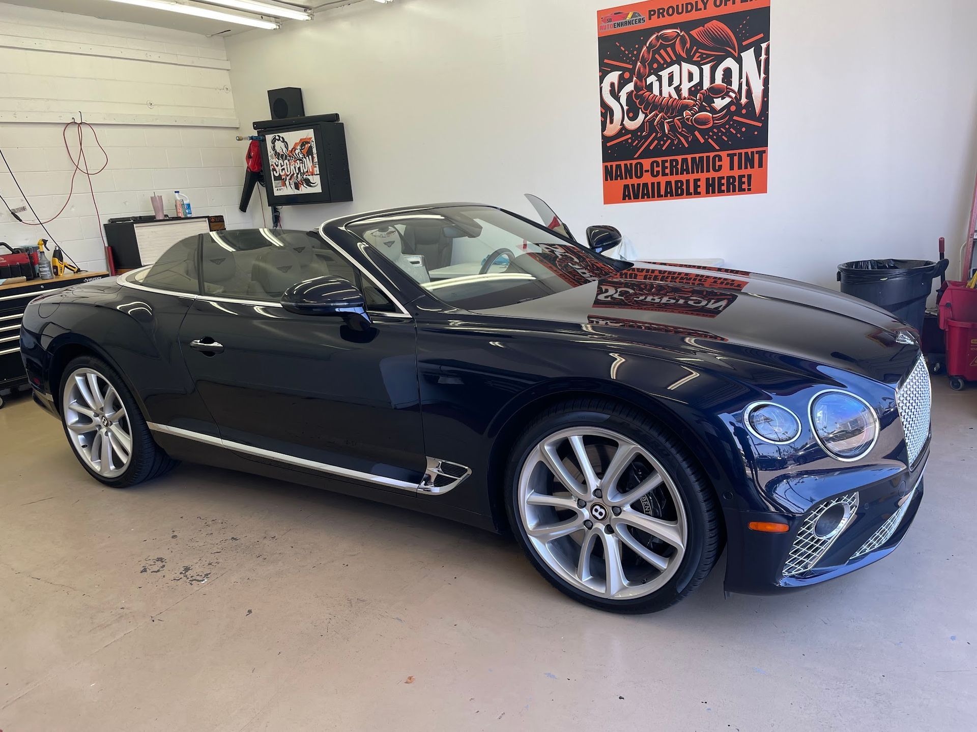 Dark blue Bentley convertible parked in a garage, with the top down. A poster hangs in the background.