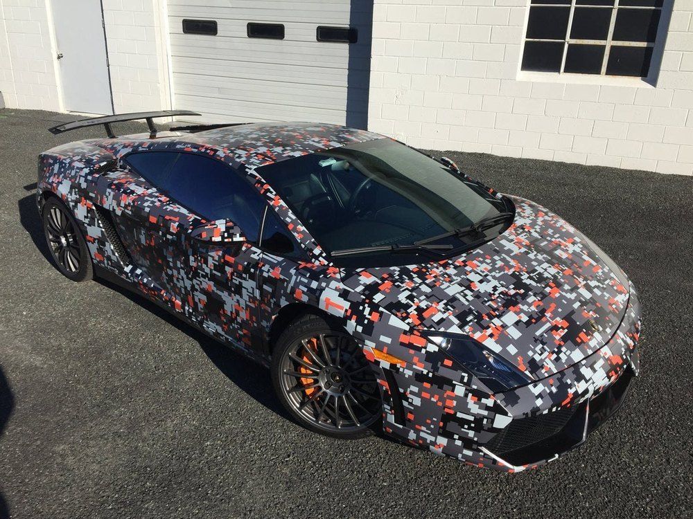 Lamborghini sports car with pixelated camouflage wrap, parked in front of a building.
