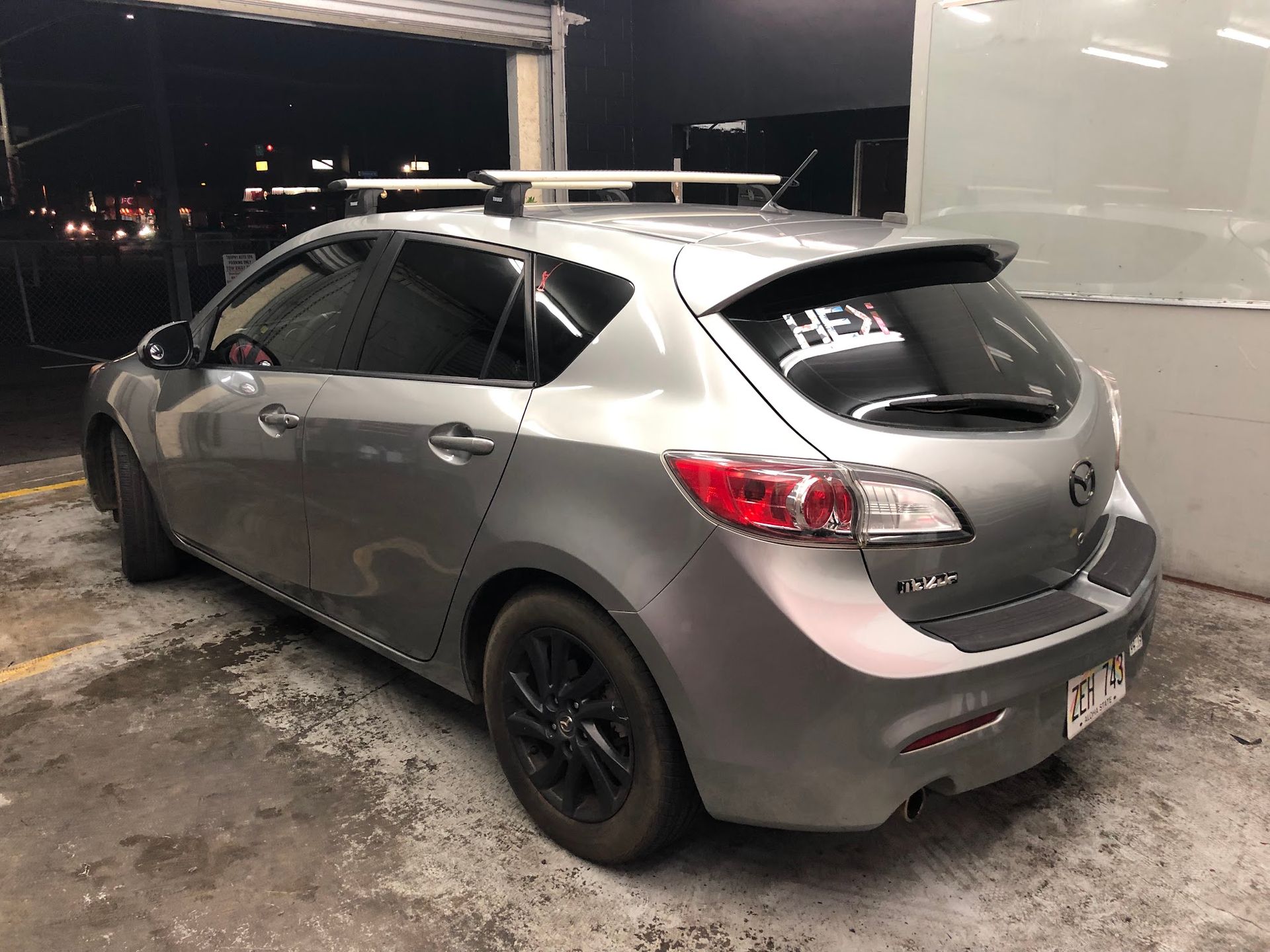 Silver Mazda 3 hatchback parked at night, black wheels, roof rack, spoiler.