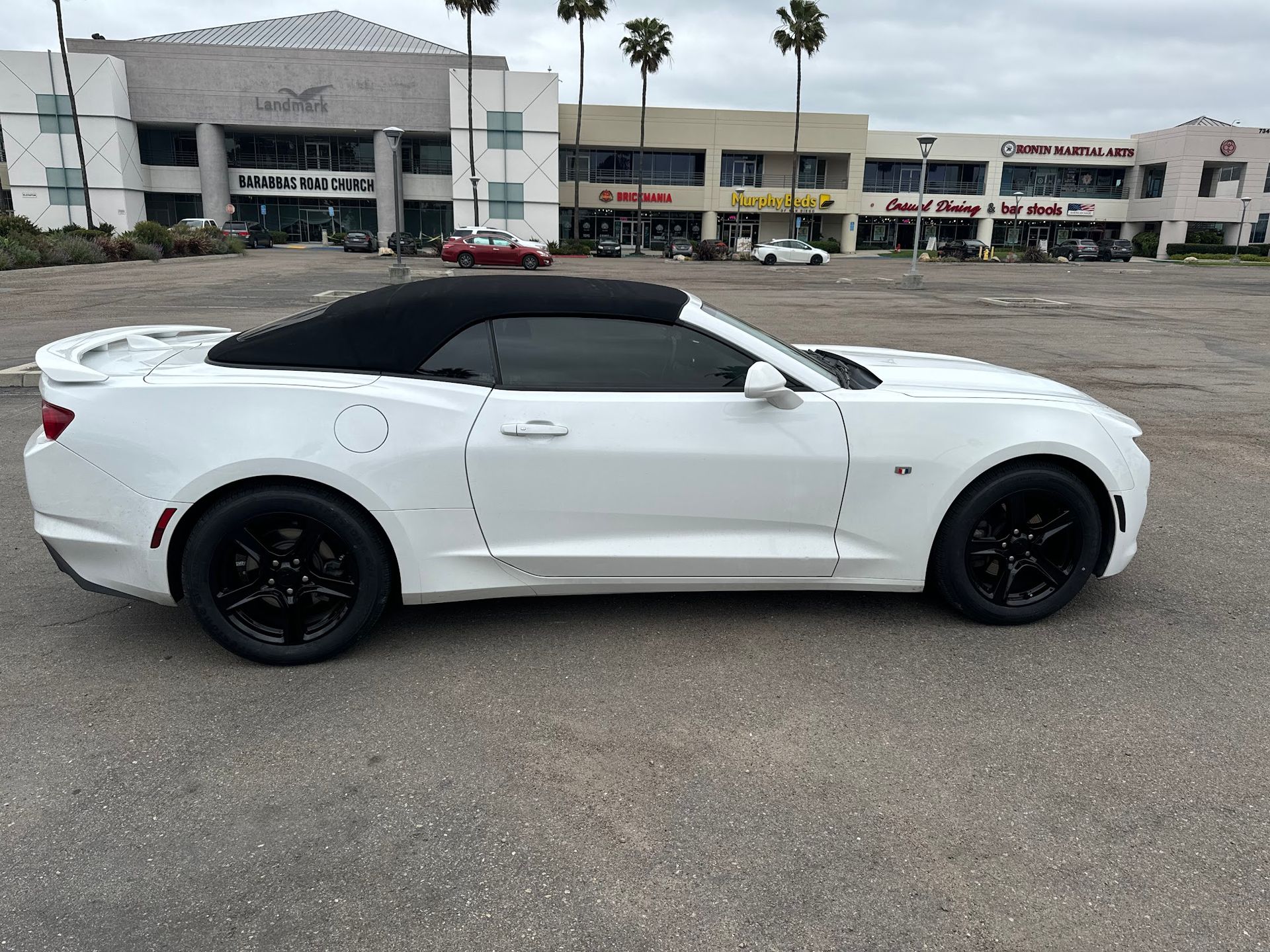 White convertible car with black wheels and top parked in a parking lot. Businesses in the background.