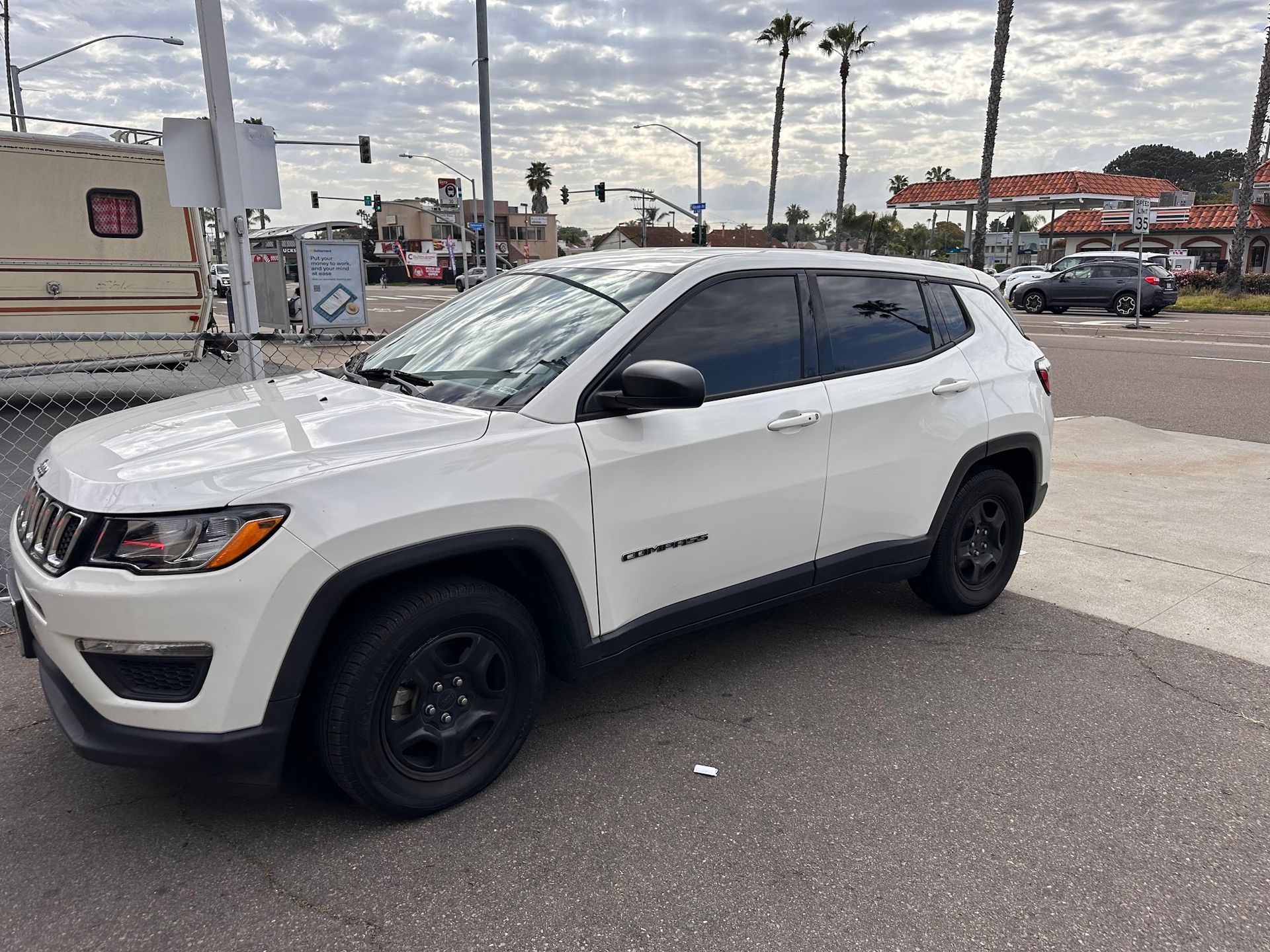White Jeep Compass on a street, parked near a building and gas station. Cloudy day.