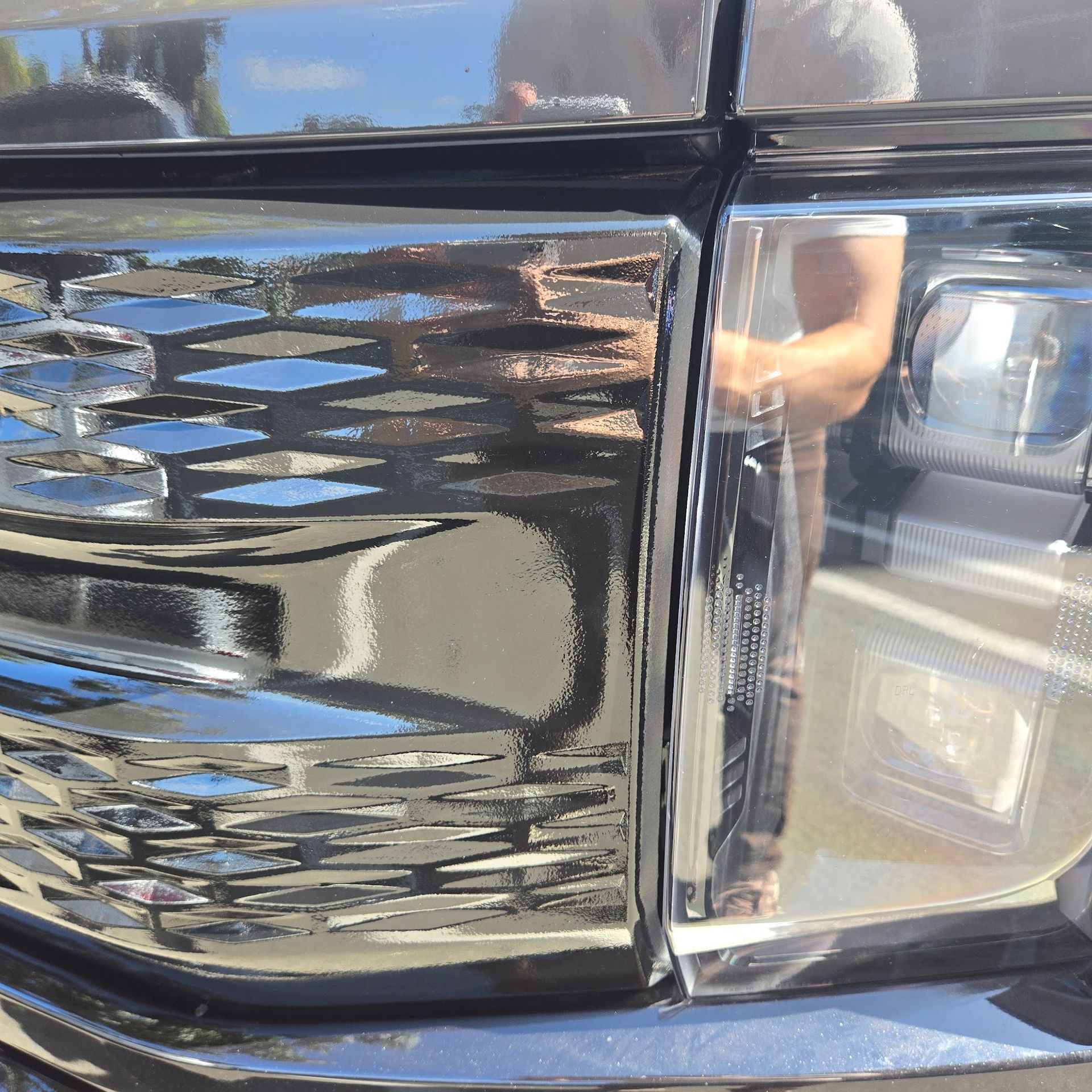 Close-up of a black truck grill and headlight, with a person reflected in the shiny surface, outdoors.