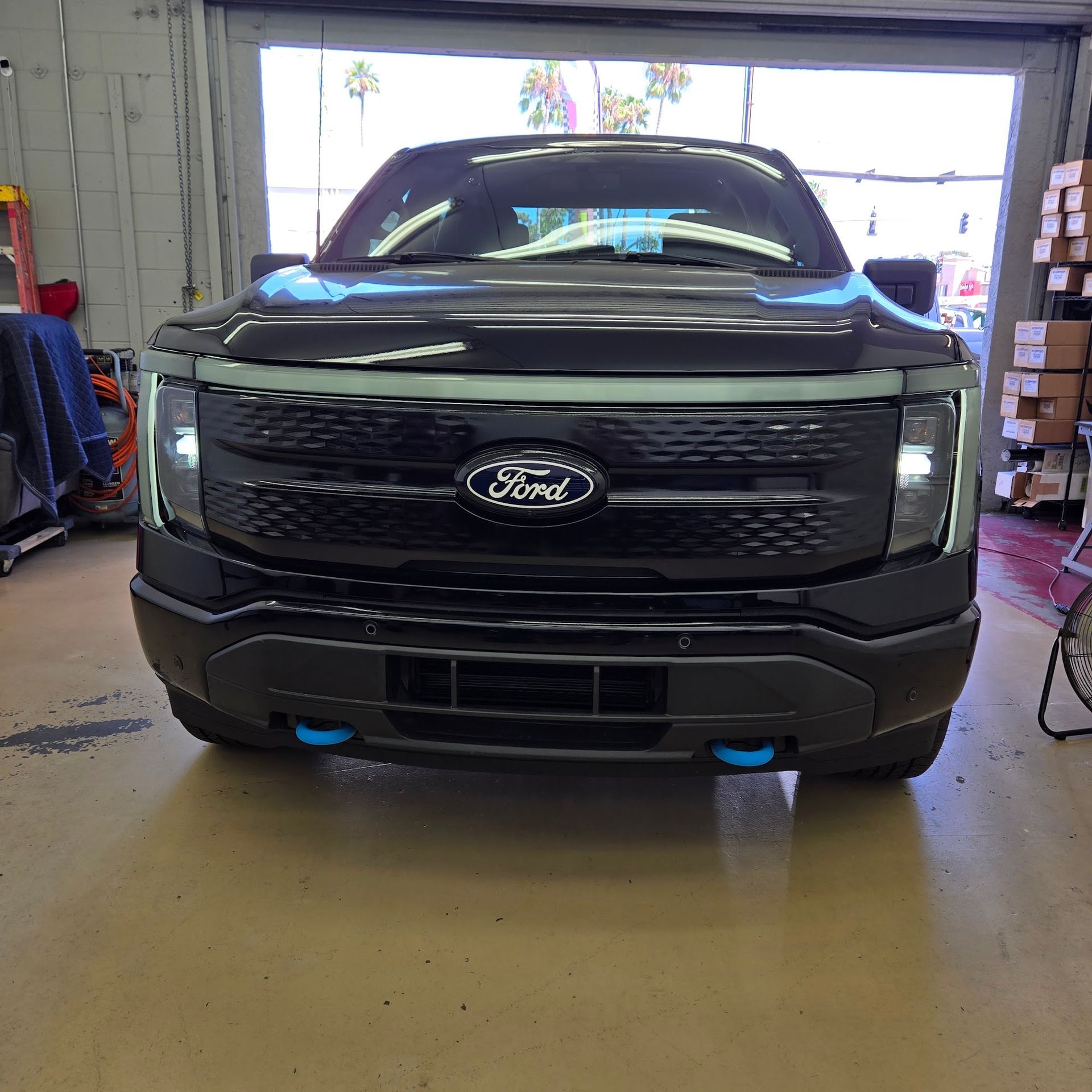 Black Ford F-150 Lightning electric truck, front view, in a garage with blue tow hooks.