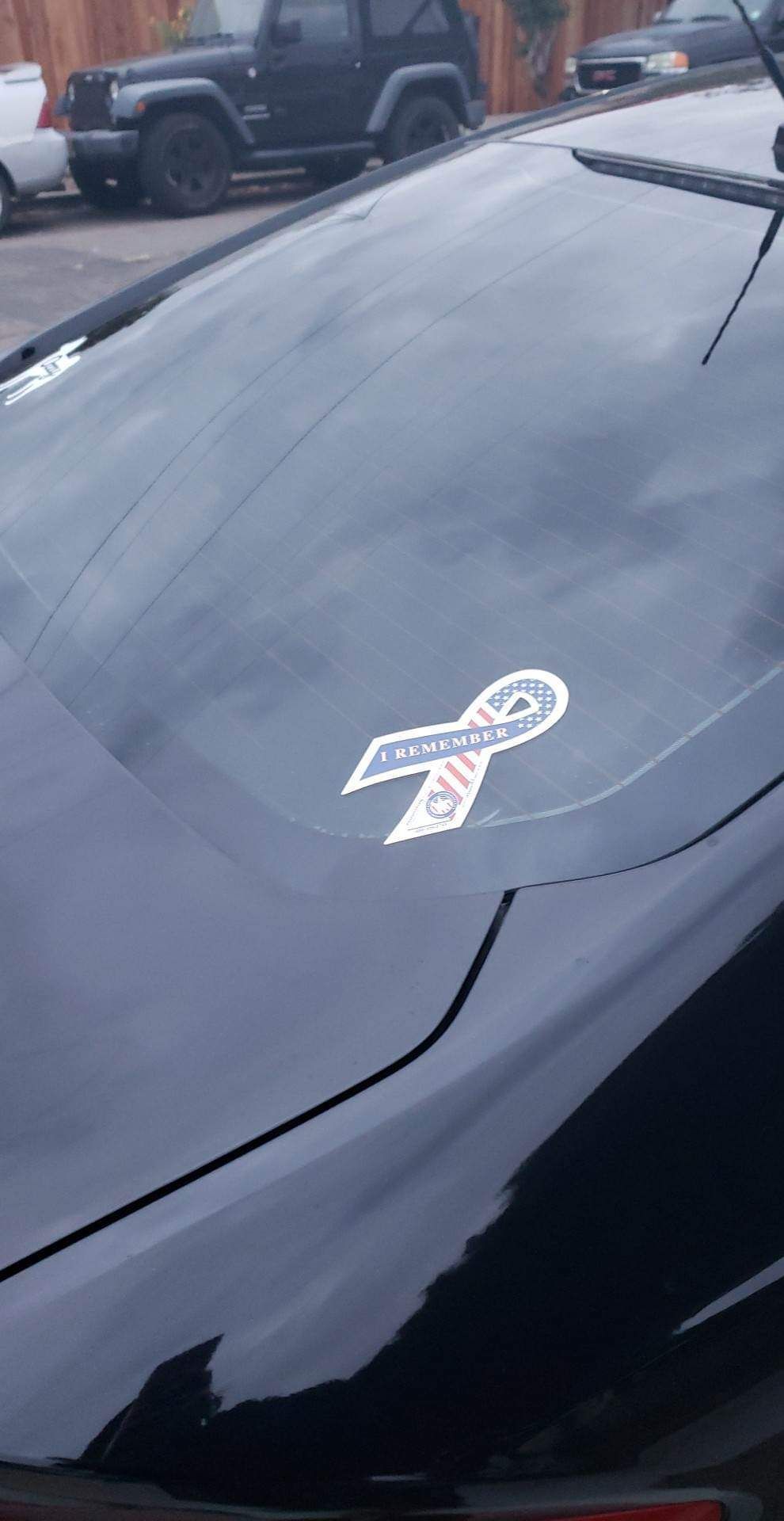 Black car with a white, blue, and red ribbon sticker on the rear window.