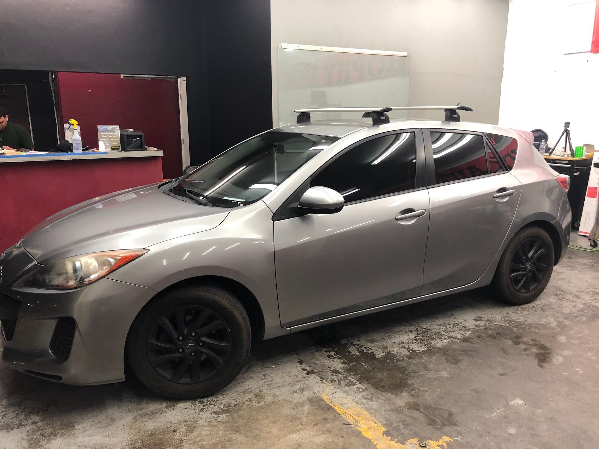 Gray Mazda hatchback with black wheels and roof rack, parked indoors.