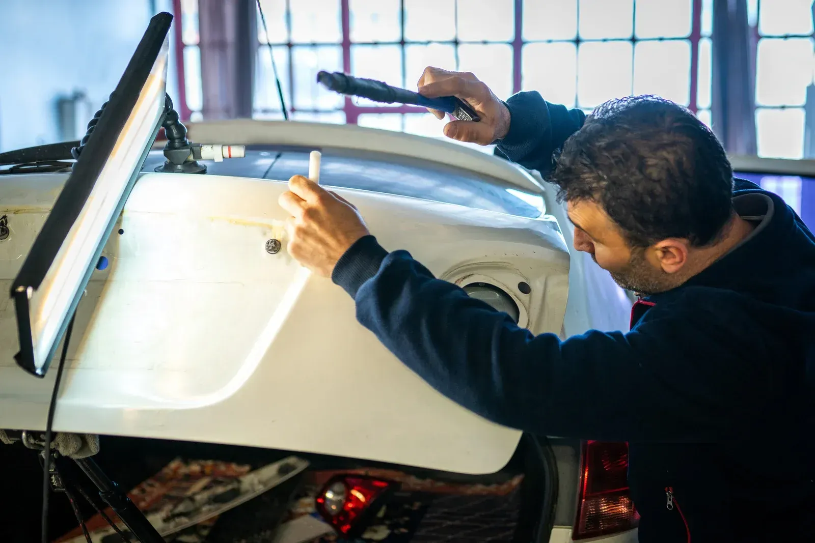Mechanic uses a light to inspect a white car for dents in a garage.