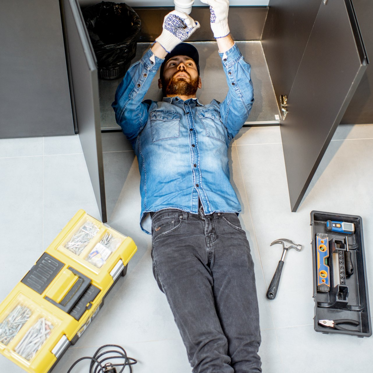 A man in a denim shirt is laying on the floor under a sink.