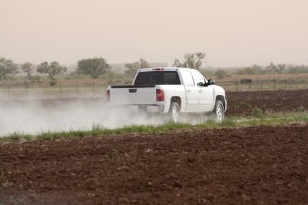 A white pickup truck driving on a dirt road, kicking up a cloud of dust through an open field.