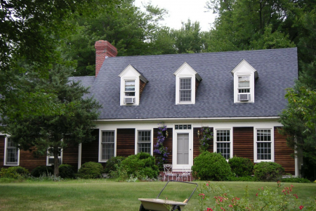 A rustic two-story house with brown wood siding, three dormer windows, and a brick chimney, set in a grassy yard.