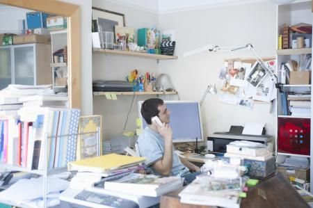 A person sitting at a cluttered desk in an office, talking on a phone while looking at a computer monitor.