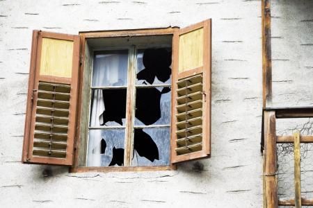 A weathered, light-colored wall with an open wooden-shuttered window containing several large, jagged broken panes.