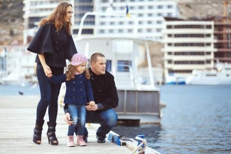 A family of three standing on a wooden pier near a boat and buildings, looking out toward the water.