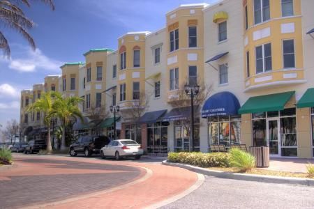 A row of multi-story buildings with storefronts, yellow accents, and awnings along a brick-paved street with parked cars.