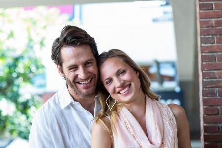A smiling couple poses closely together outdoors in front of a brick wall and blurred greenery.