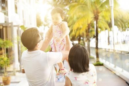 A person lifts a toddler into the air on a sunny outdoor patio, while another person watches nearby.