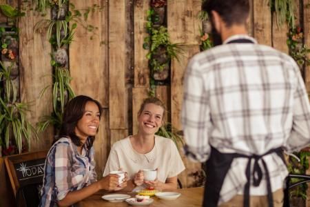 A waiter in a plaid shirt and apron stands by a table as two smiling customers hold cups in a cafe with a plant wall.