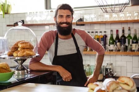 A barista wearing a black apron and a two-toned shirt smiles while standing behind a counter with pastries in a cafe.