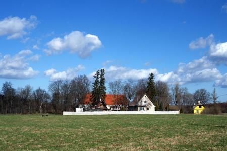 A rural landscape featuring a white-walled compound with orange and white buildings behind a grassy field under a blue sky.