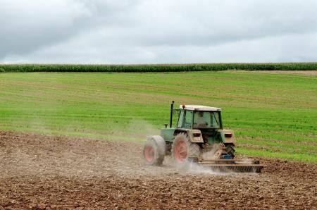 A green tractor tills a field of dark brown soil against a backdrop of green grass and a cloudy sky.