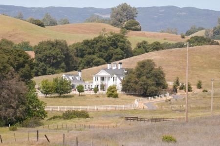 A white colonial-style house sits in a valley surrounded by rolling, grassy hills and scattered oak trees.