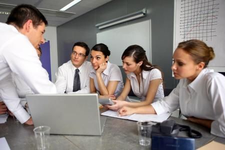 A group of people in business attire working together around a laptop in an office meeting room.
