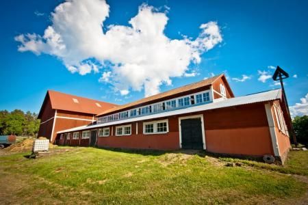 A long, red barn with white-trimmed windows and a rust-colored roof set against a bright blue sky with fluffy white clouds.