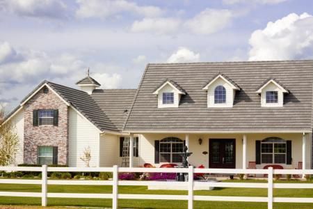 A cream-colored farmhouse with a grey shingle roof, three front dormers, brick accents, and a white rail fence.