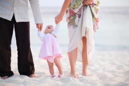 A toddler in a white cardigan and pink dress held by two adults on a sandy beach.