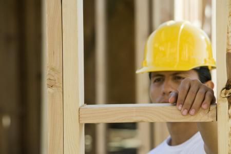 A construction worker wearing a yellow hard hat looking through the wooden frame of a building under construction.
