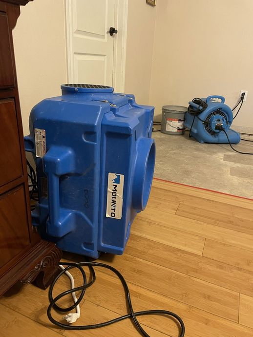 Blue dehumidifier on hardwood floor with power cord, near a rug and a blue blower in a room