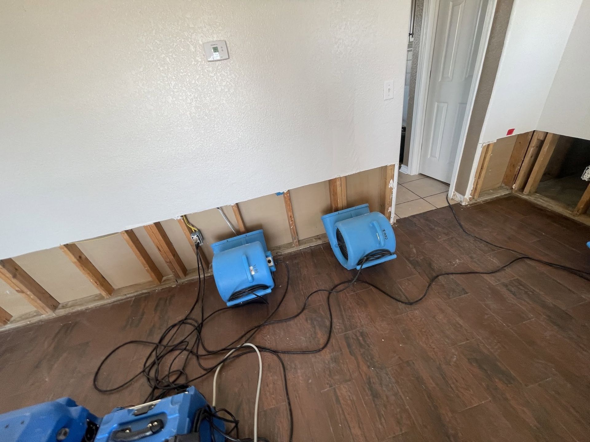 Two blue floor fans on a hardwood floor against a wall with exposed trim and cords.