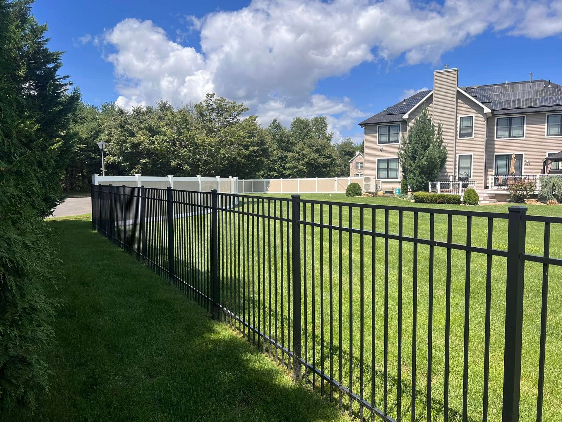 A black fence surrounds a lush green field in front of a large house.