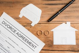 A document titled Divorce Agreement, a pen, two wedding rings, and paper cutouts of a house and car on a wooden table.
