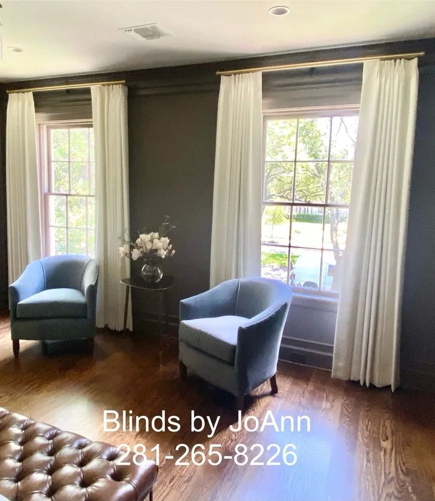 Two blue chairs face windows with white drapes. Dark walls, gold curtain rods, and a round table.