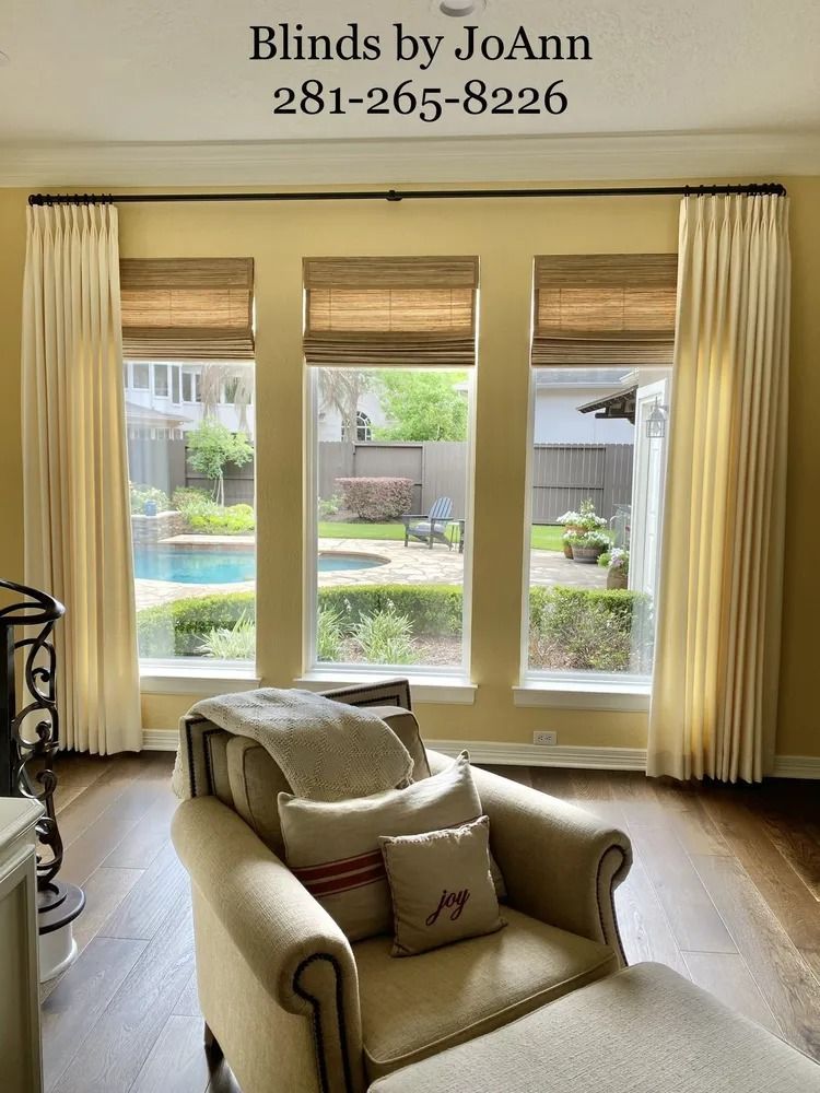 Living room with window treatments: pleated curtains, woven shades. Chair with pillows, view of pool.
