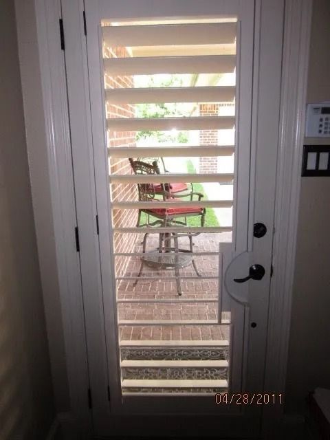 White door with open shutters, revealing a brick patio with outdoor furniture.