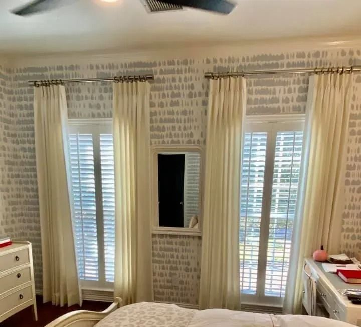 Bedroom with white curtains, shutters, and patterned wallpaper. Mirror between windows.