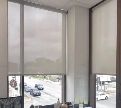 Window with neutral-colored roller shades, overlooking a street with cars and buildings.