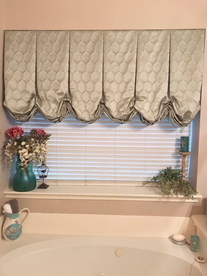 Pale green fabric valance above a window with blinds. A teal vase holds flowers, and a candle sits on the windowsill.