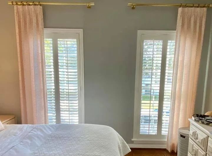 Two windows with white shutters, pink curtains, and gold rods in a bedroom with a gray wall.