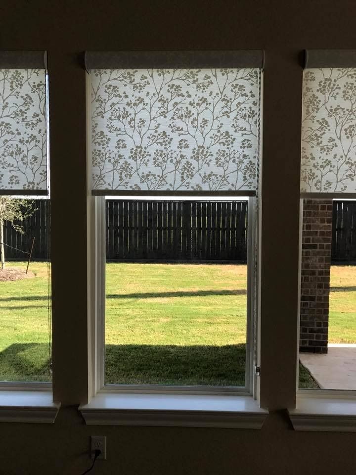 Three windows with floral print roller shades, overlooking a grassy yard and a dark wooden fence.