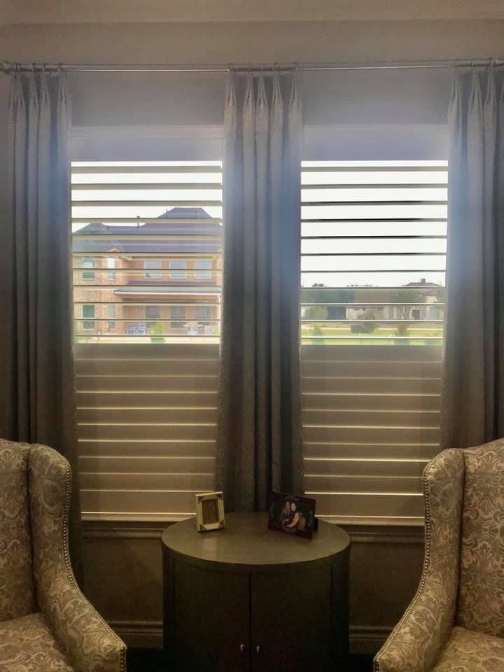 Two windows with gray curtains, shutters, and a round table between patterned armchairs.
