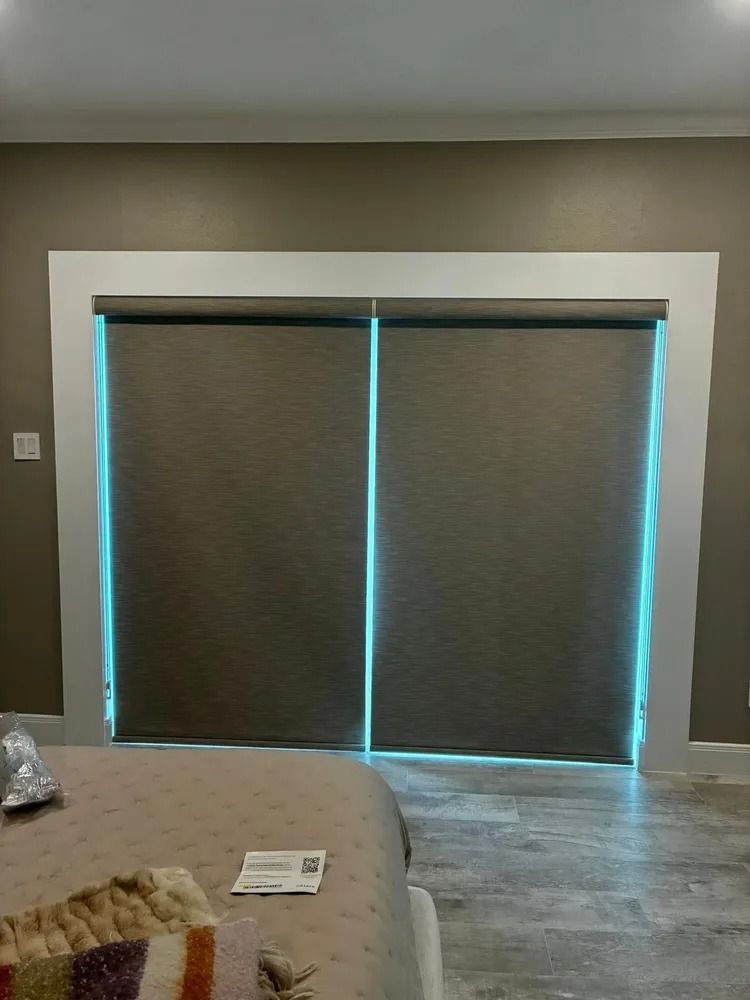 Two closed gray window shades within a white-framed doorway, casting a blue glow on light-gray walls, above a patterned bed.