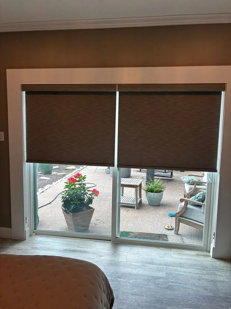 Two closed brown roller shades over a glass door with an outdoor patio visible.