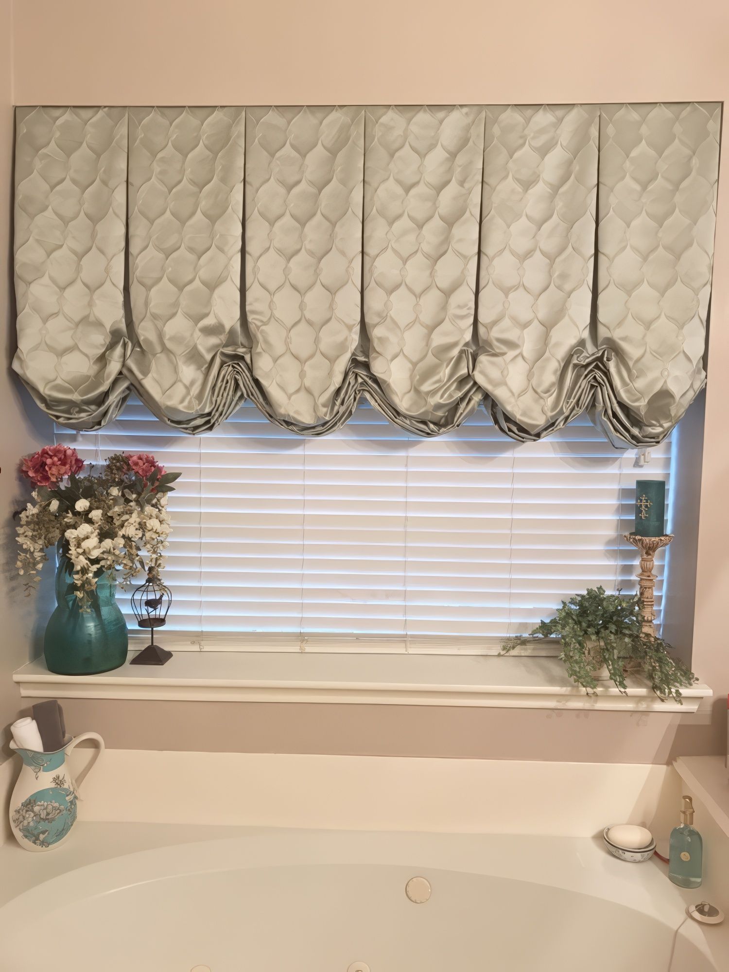 Window with draped valance and closed blinds; floral arrangement on the windowsill, a bathtub below.