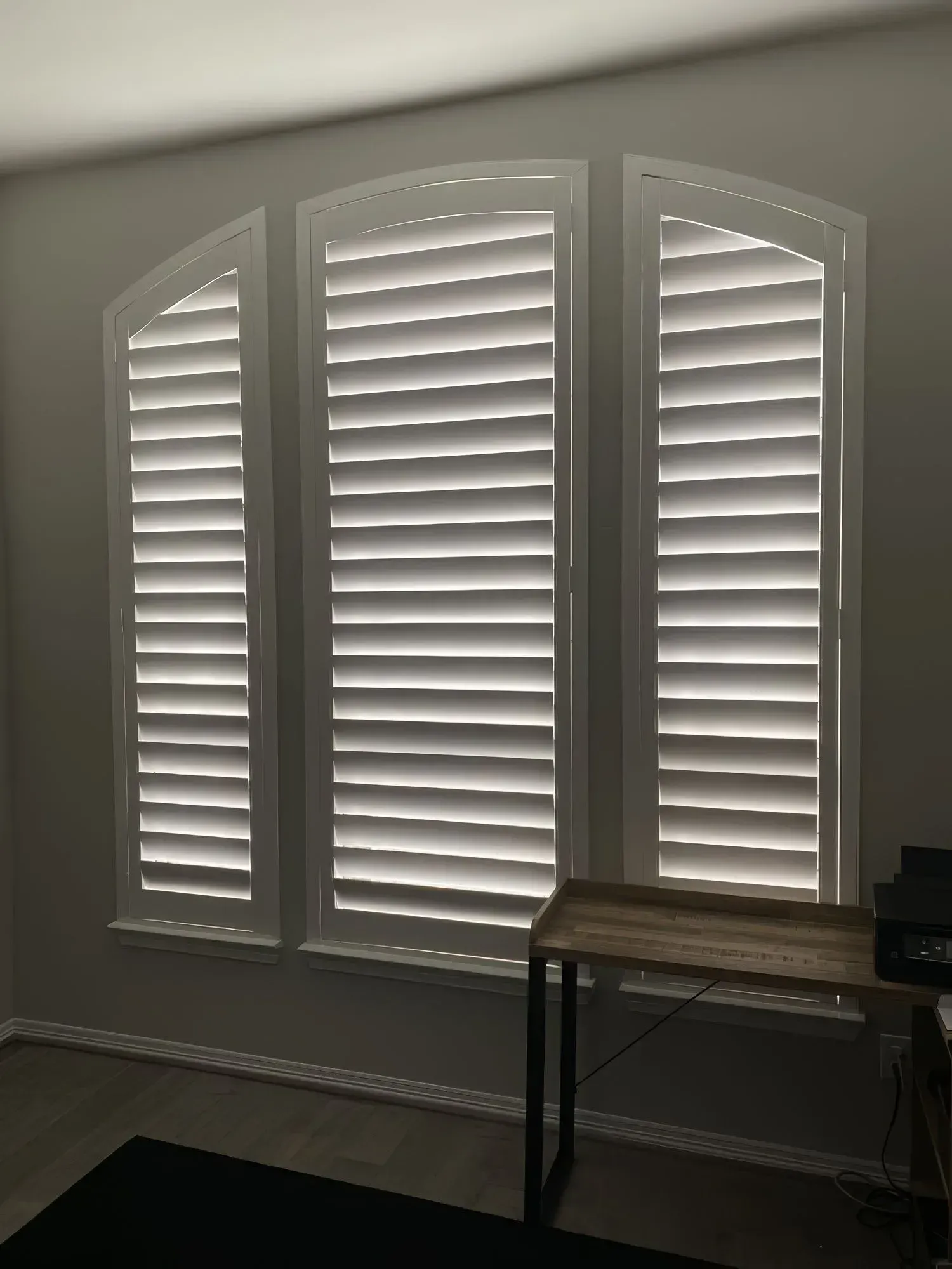 White shutters cover an arched window, casting horizontal light and shadow in a room with a desk.