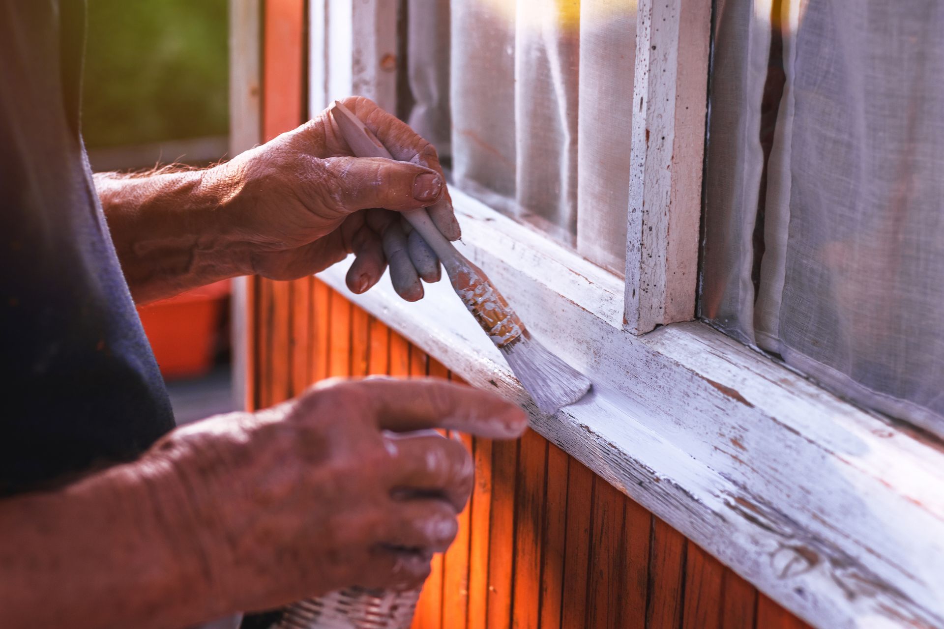 Person painting white paint on a wooden window frame outdoors.