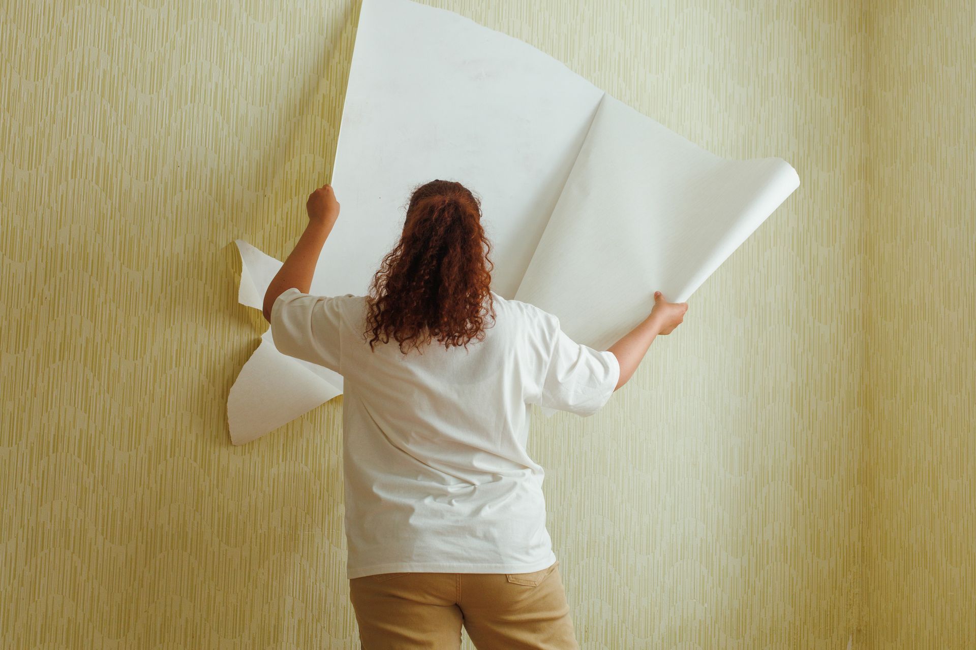 Woman peeling off wallpaper from a textured yellow wall, back view.
