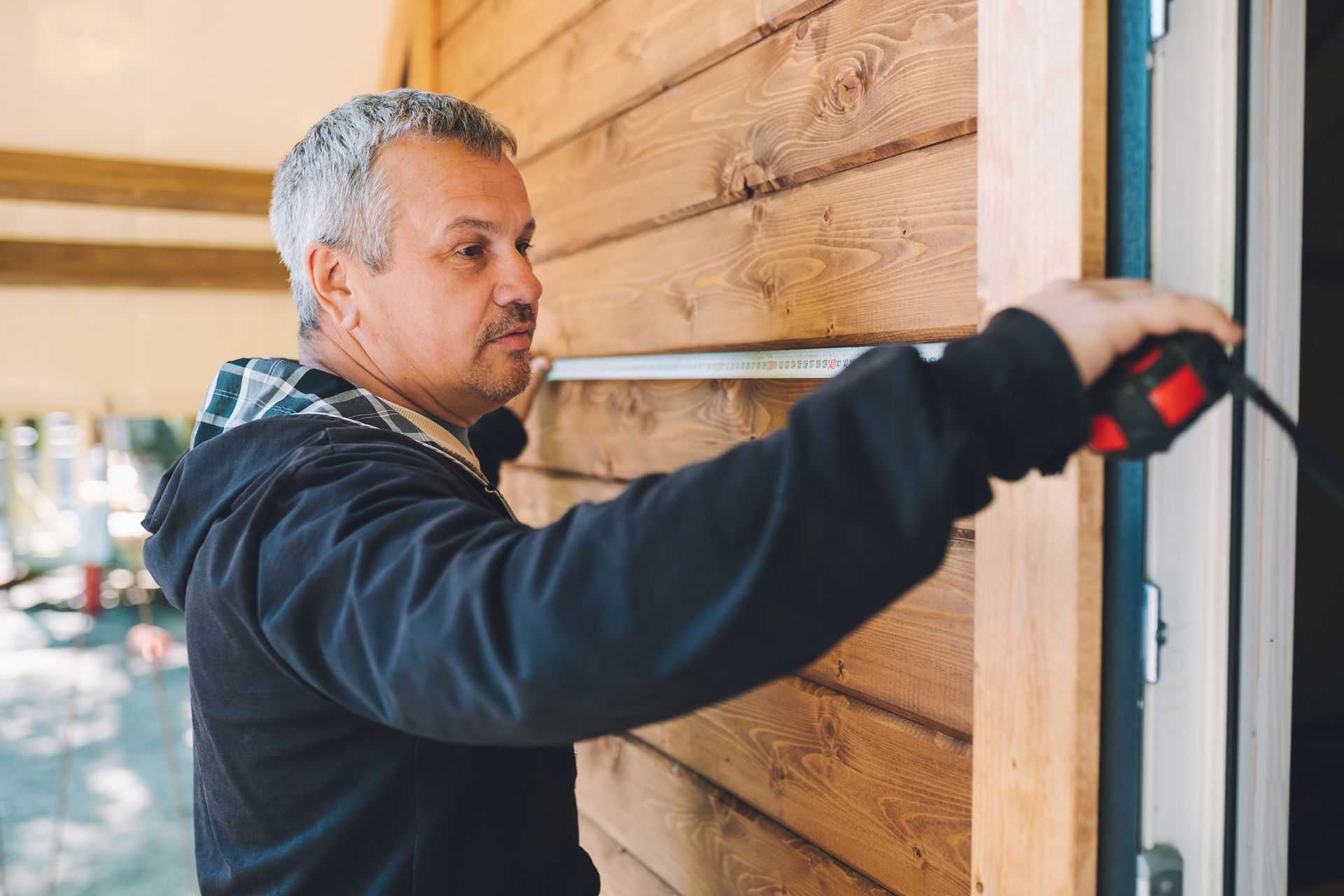Man with gray hair, measuring wooden door frame with a red tape measure outside a building.