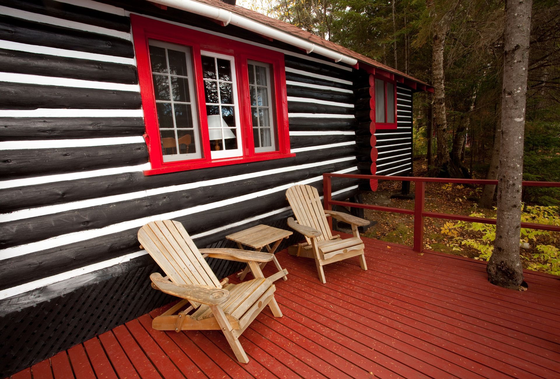 Cabin with black and white logs, red trim and deck, two wooden chairs.